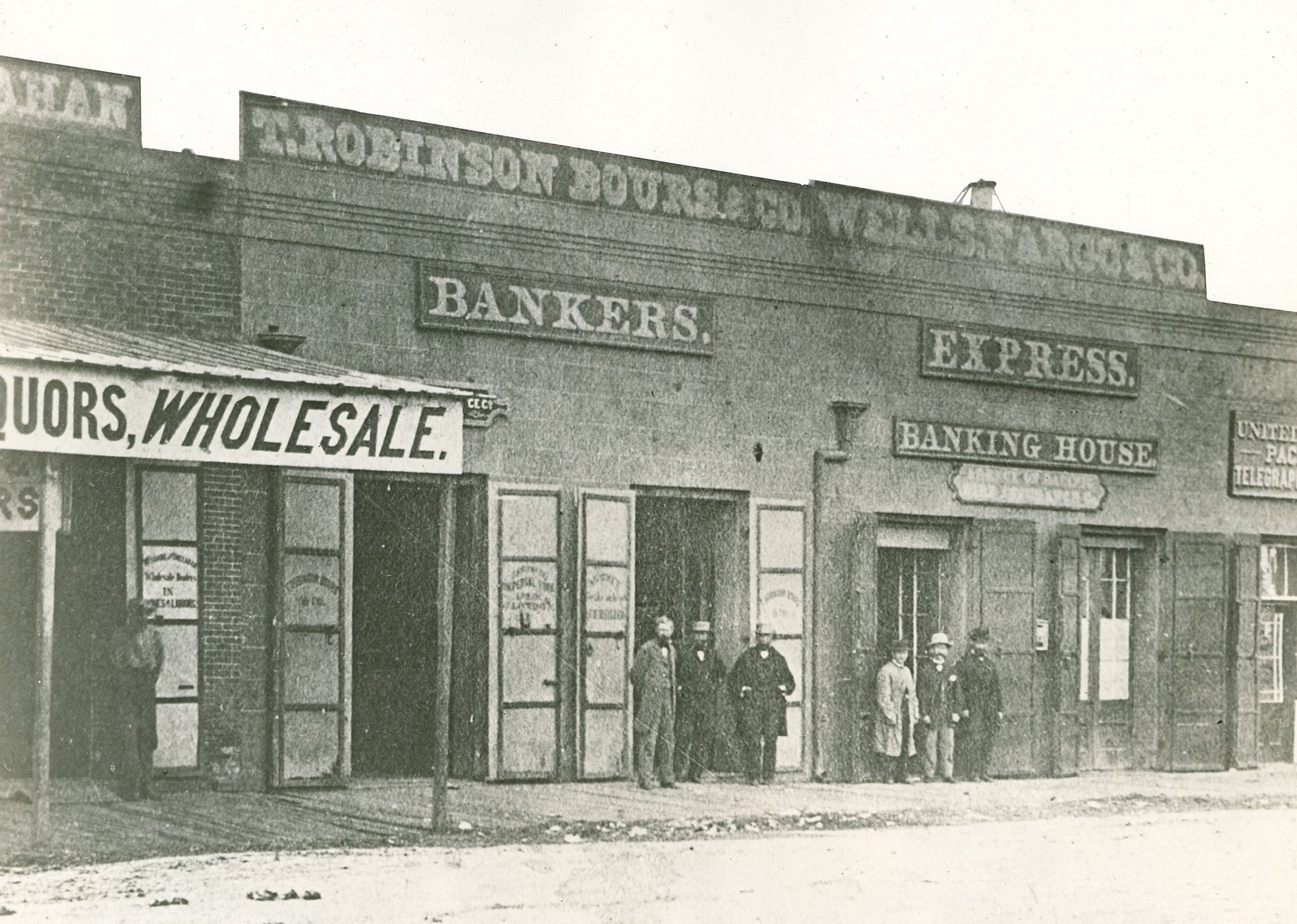 A black and white image shows several single-story commercial buildings in a row. Signs read T. Robinson Bours & Co Bankers and Wells, Fargo & Co. Express and Banking House. Seven men are on the sidewalk in front of the bank buildings.