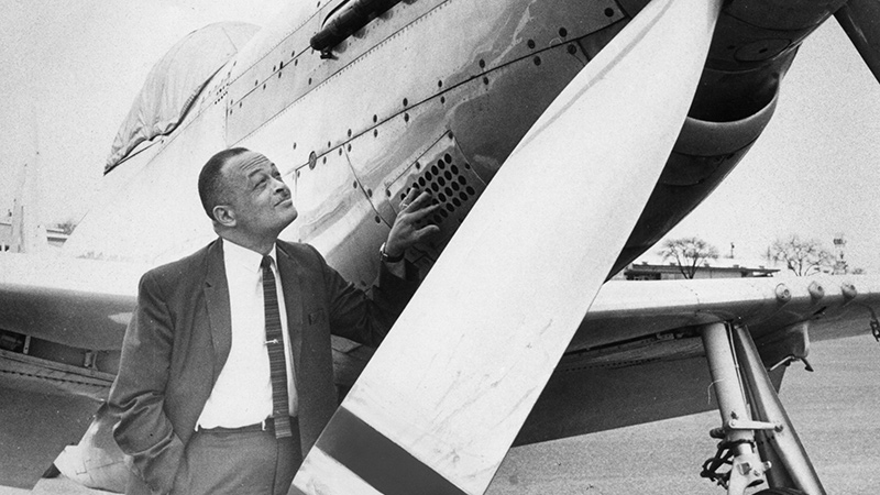 Banker George Roberts returns to an airfield to reminisce. He is wearing a business suit with white collared shirt and tie. He stands beside an airplane, gazing reflectively. Historic black and white photograph.