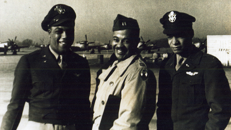 Three men in WWII era Army Air Corps. uniforms stand outside on a field. The man on the left and the man on the right have dark coats. The man in the middle is wearing a tan coat.