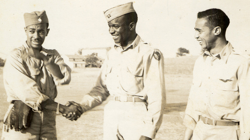 Three men in WWII era Army Air Corps. Uniforms stand outside on a field. The man on the left and the man in the middle are shaking hands.