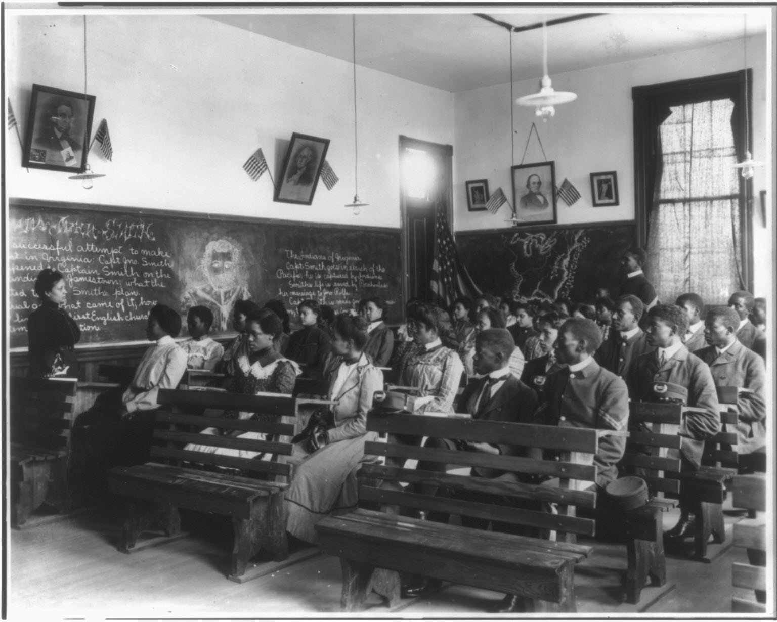 Black and white photograph of a class of young students watching a teacher who stands in front of a blackboard.