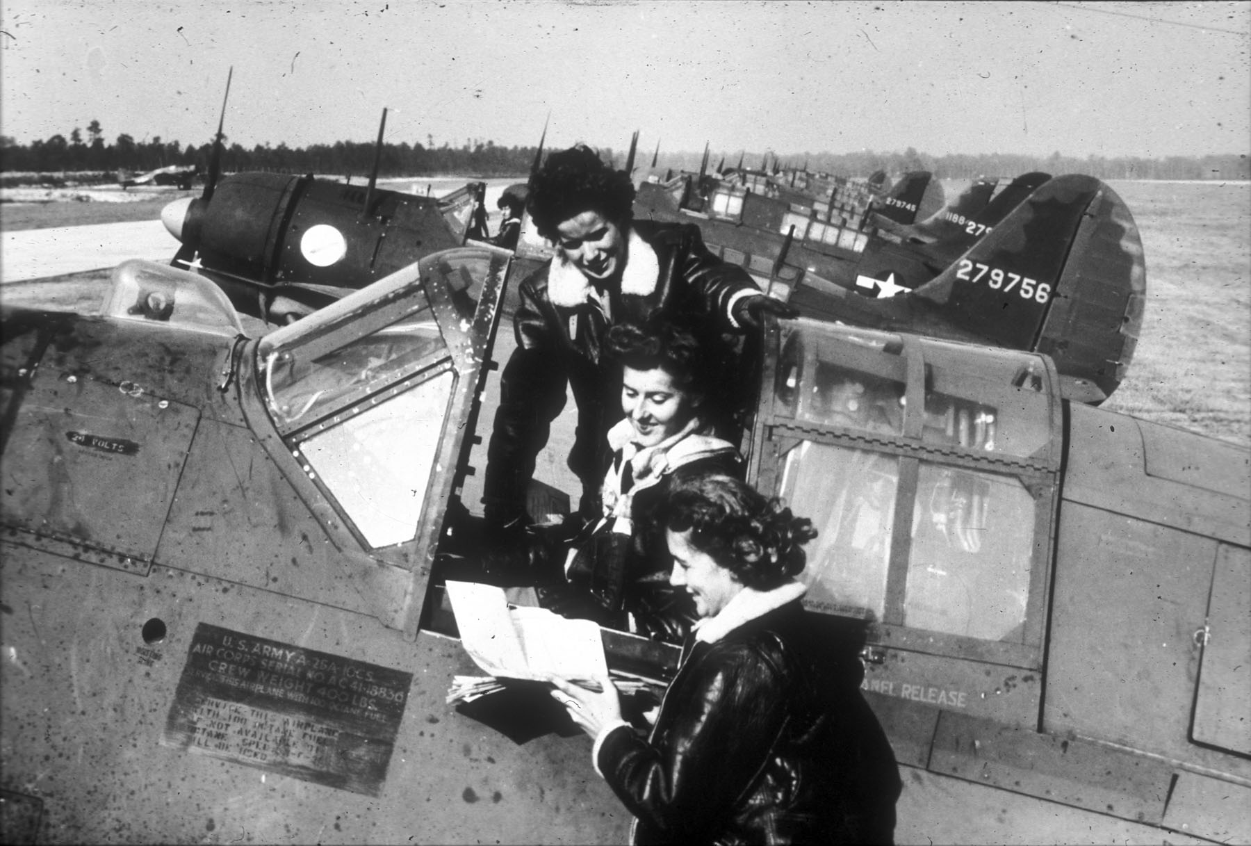 Three women at the cockpit of a B-17 Bomber. One sits in the pilot’s seat while the other two stand on either side. All three women are looking at an opened manual and are wearing flight bomber jackets.