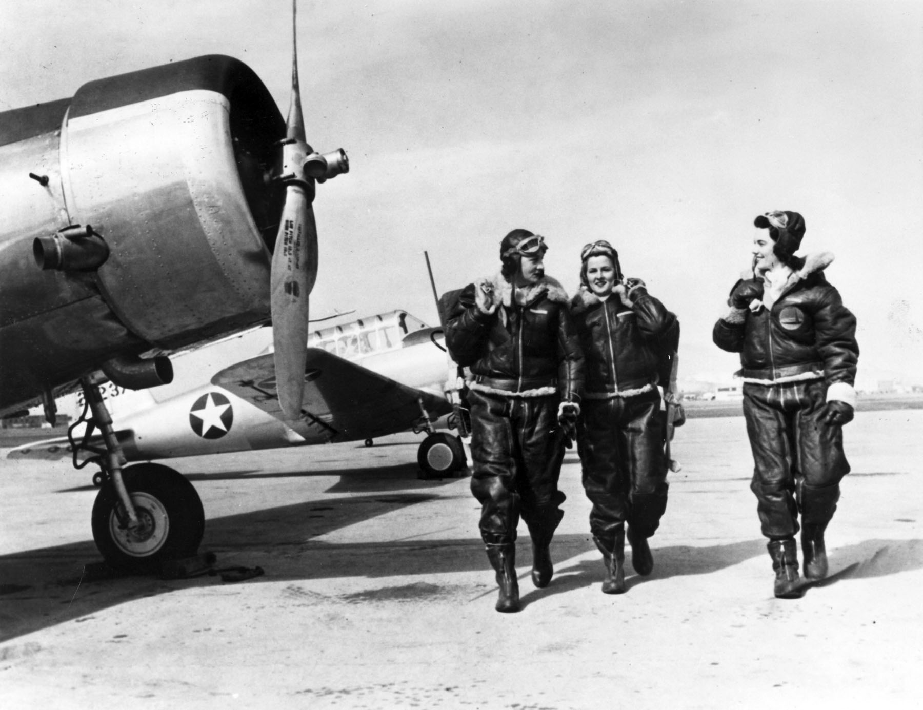 Three women wearing World War Two era pilot gear walk on a tarmac, with two parked planes on the right.