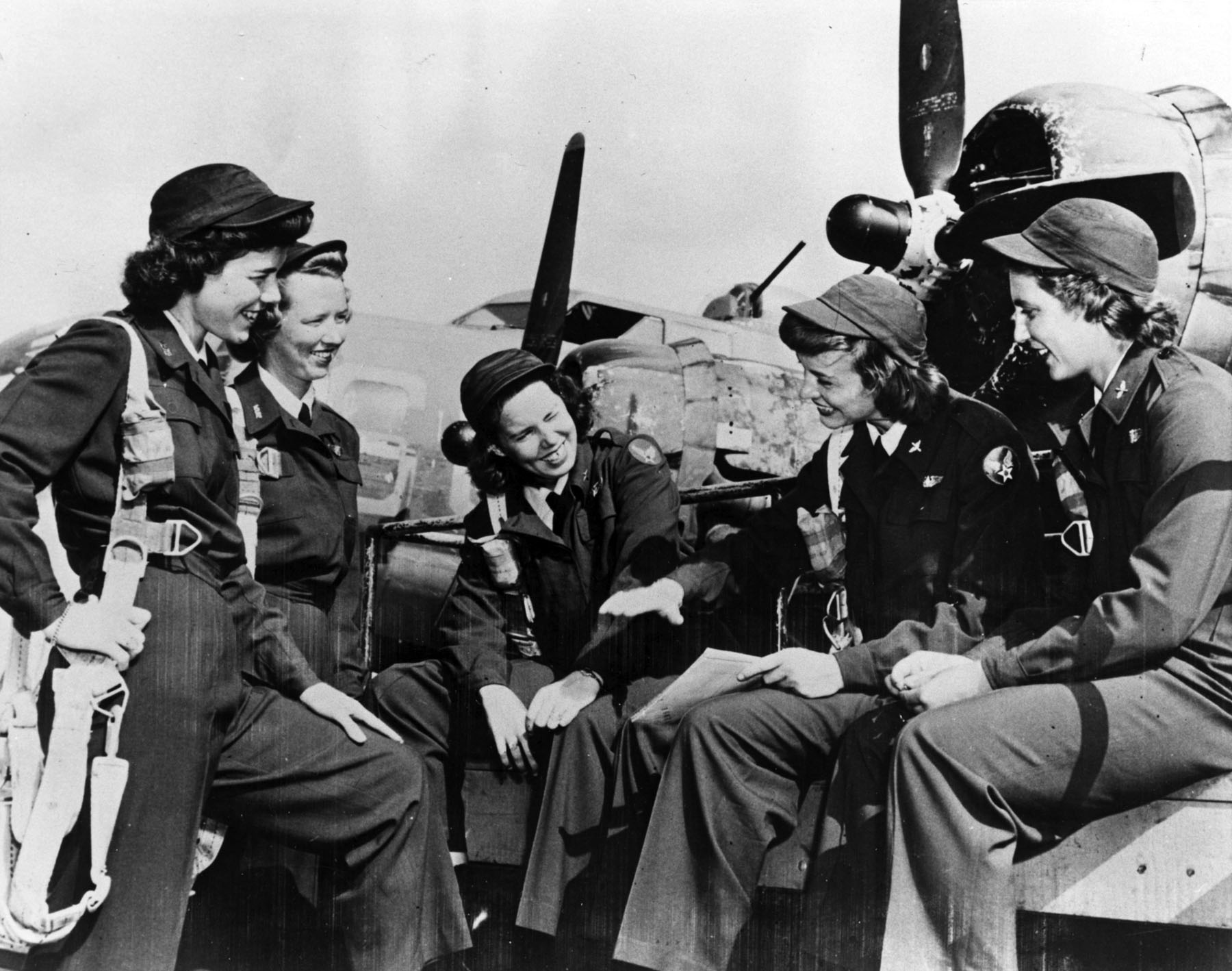 Five women dressed in WWII era uniforms gathered on an airfield with two military planes visible in the background.