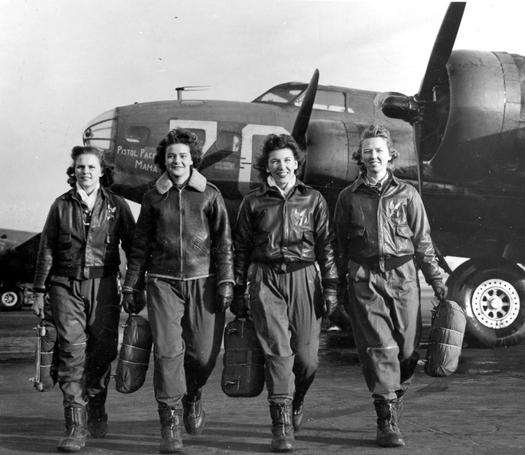 Four women wearing WWII era flight bomber jackets walk side by side away from a B-17 bomber plane parked behind them.