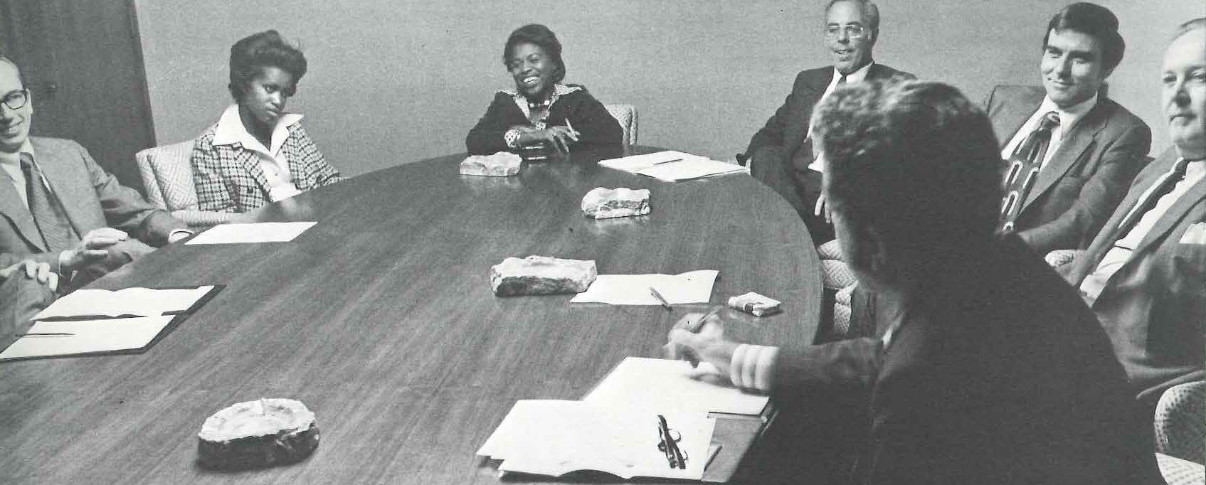 A group of men and women sit around an oval table listening while Fernando Guzman speaks.