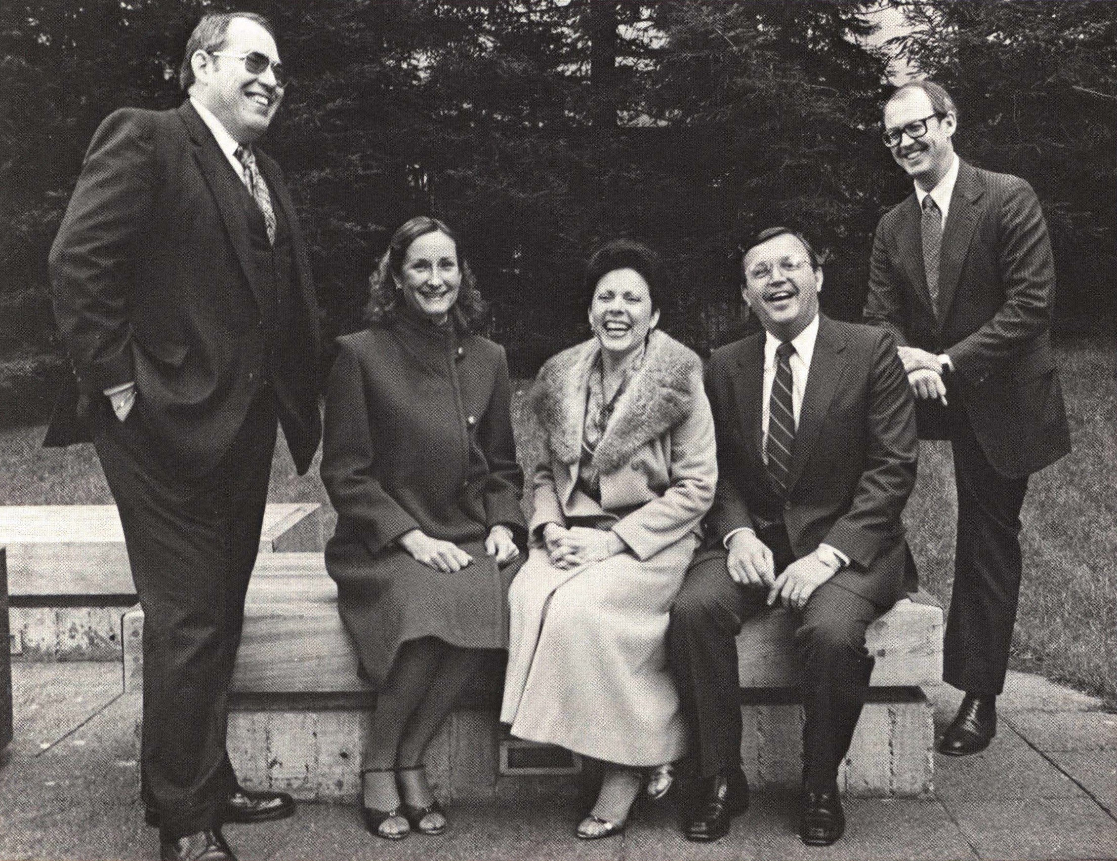 Black and white photograph featuring 5 people outside. Two stand and three sit on a concrete bench.