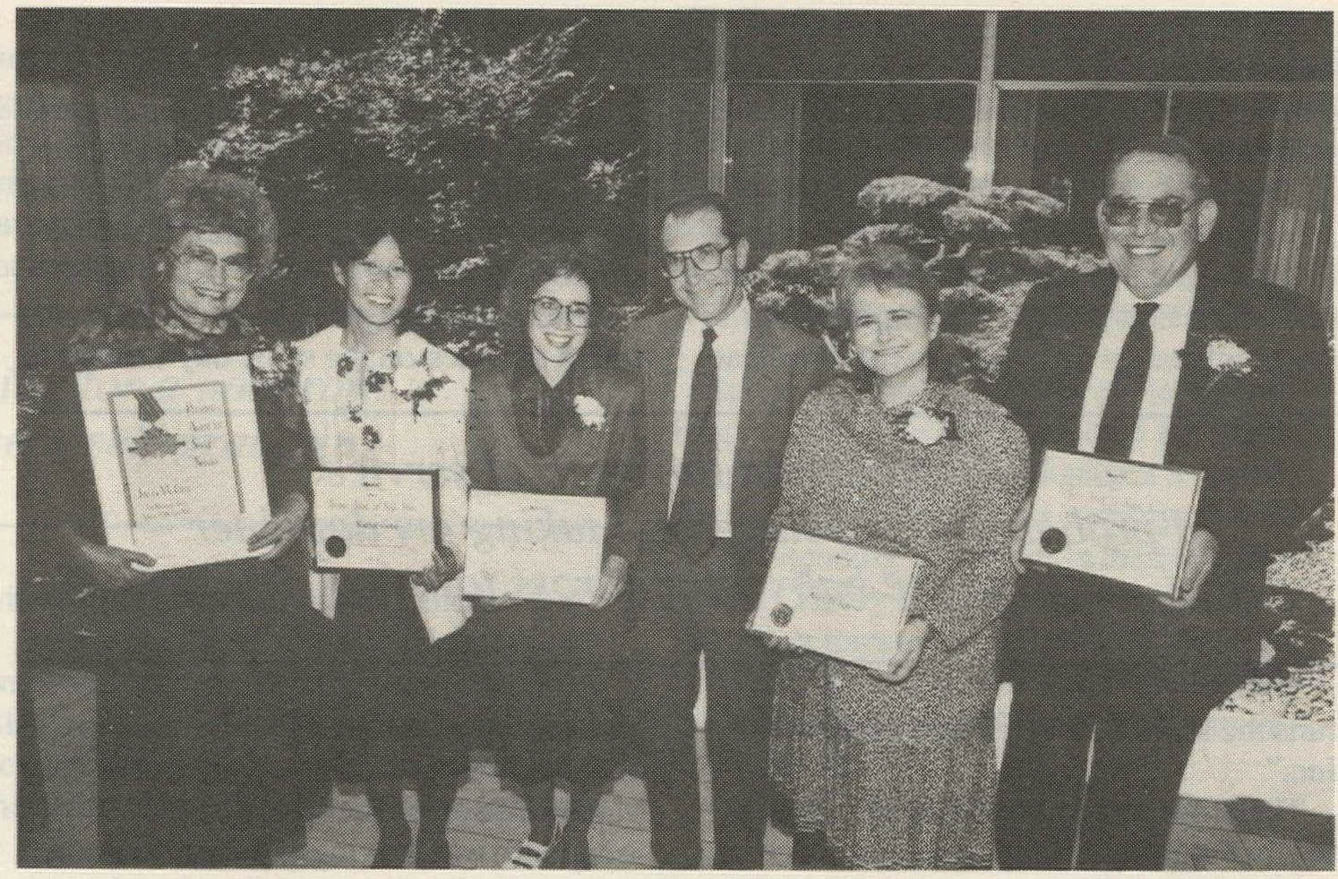 Six people in business attire stand in a line facing the camera. Four are women, two are men. Five of the people are holding a framed certificate, four are identical. The woman on the left most holds a unique certificate that is bigger and more elaborate.