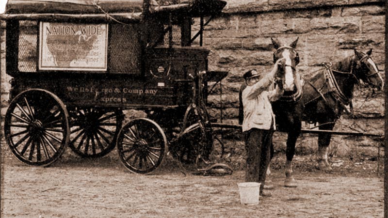 A Wells Fargo wagon is parked before a brick wall. Hitched to the wagon are two black horses with white face stripes. An employee with a bucket of water and a sponge is wiping down one horses face.