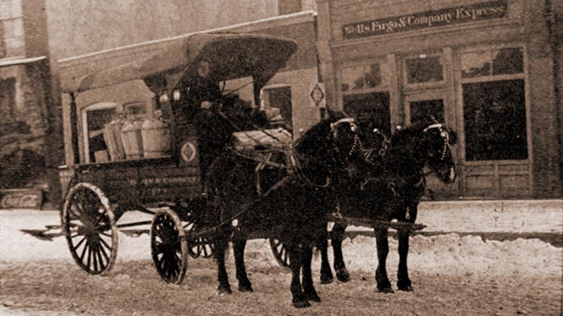 A street scene with buildings in background and a Wells Fargo wagon pulled by two horses in foreground. Each black horse is wearing a raincoat to keep warm and dry.