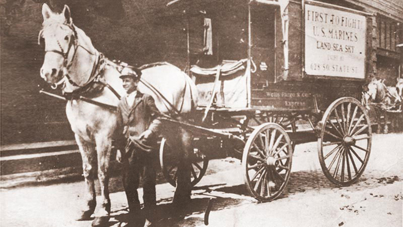 Wells Fargo express wagon pulled by one large gray horse. Young man stands at horse’s left side. Banner on wagon reads in large letters First to Fight U.S. Marines Land Sea Sky Enlist at 628 South State Street.