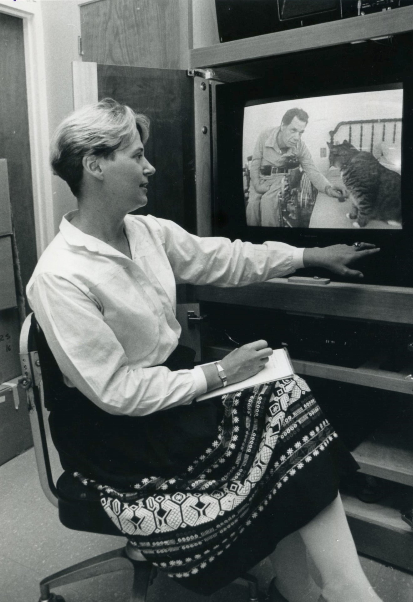 A woman wearing a long skirt and white blouse is seated in a chair before a TV. On the screen a man is petting a cat.