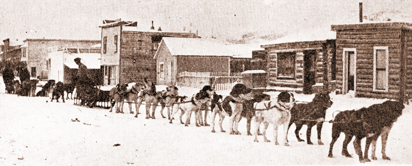 A snowy landscape with small wood buildings in the background. A team of dogs stands ready as a man loads items onto a sled.