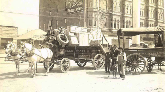 A street scene with a fully loaded Wells Fargo wagon on the left and three men standing to the right in front of a smaller buggy. One man in overalls holds a large hound dog with a leash. Image link will enlarge image.