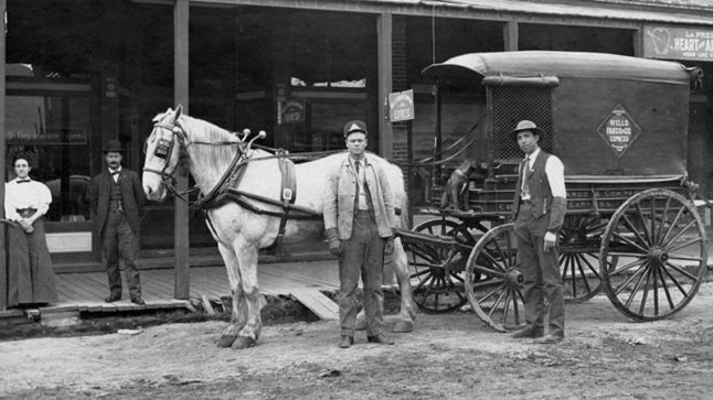 A street scene with three men and a woman standing beside a Wells, Fargo wagon being pulled by one white horse. Sitting in the driver’s seat is a small dark dog. Image link will enlarge image.