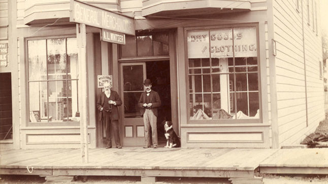Two men in suits stand an entrance. A border collie sits at attention beside them. The sidewalk out front is constructed of wood and a sign in the window next to the Wells Fargo office reads: Dry Goods & Clothing. Image link will enlarge image.