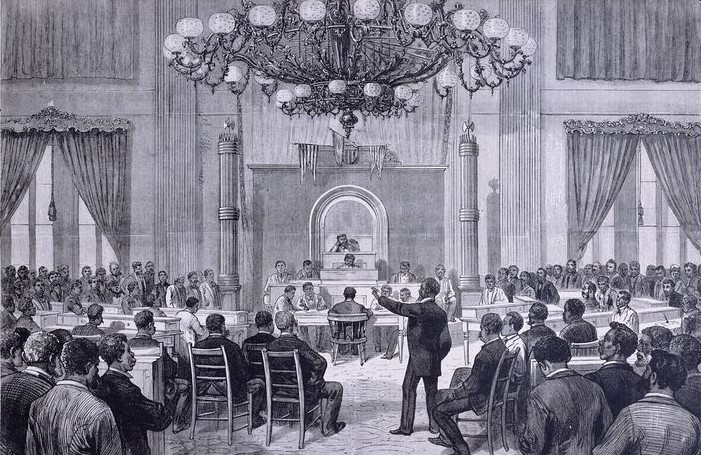 A black and white drawing shows a group of approximately 50 men sitting and standing in a large, richly decorated assembly room. They face a panel of several men seated at the center of the room and presiding over the scene.