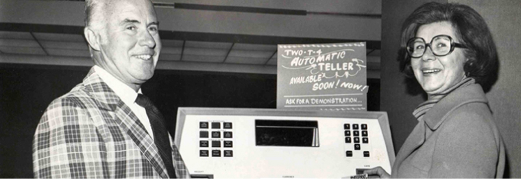 A black and white image of a man and a woman standing before an early version of an ATM machine. A sign above the machine reads: Automatic Teller Available Soon Ask for a Demonstration.