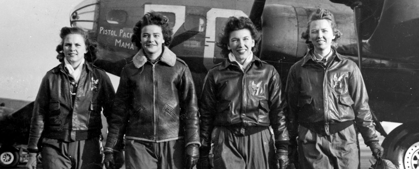 Four women wearing World War Two era flight bomber jackets walk side by side away from a B-17 bomber plane parked behind them.