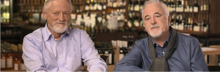 Two men are seated at a table. The man on the left has grey hair and beard and is wearing a lavender shirt. The man on the right has grey hair and goatee and is wearing a blue shirt and black scarf. Behind them are visible shelves of wine bottles.