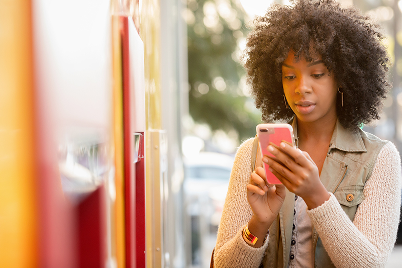 Woman with long dark hair wearing denim jacket looks at phone while walking away from Wells Fargo ATM. Color image
