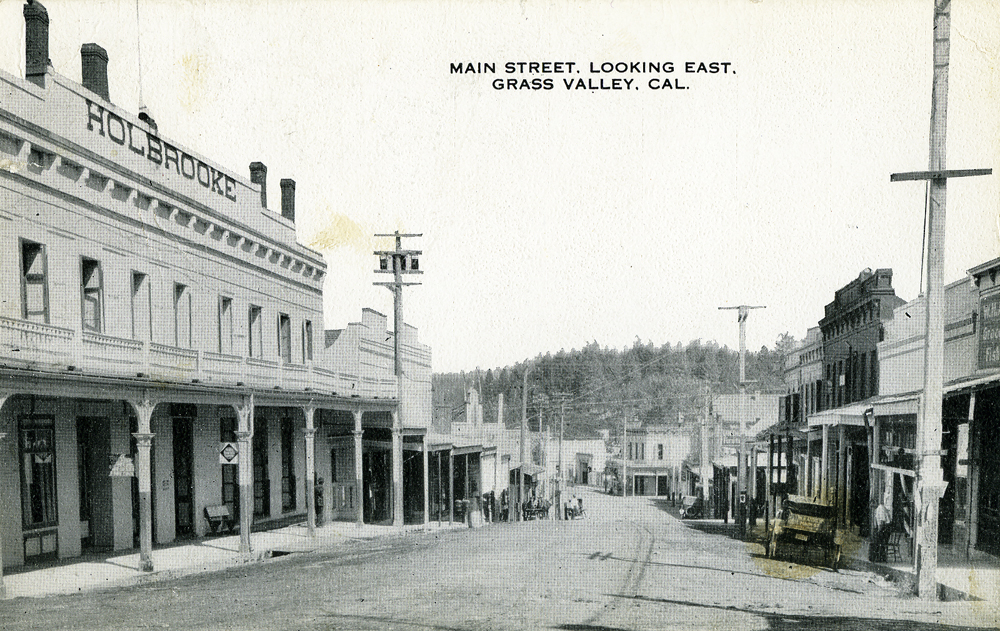 A postcard of a street scene with wooden buildings on either side and wood sidewalks. In the distance large pine trees rise over a hill. Signage on the largest building on the left reads: Holbrooke. Top of card reads: Main Street. Looking East. Grass Valley, CAL. Image link enlarges image.