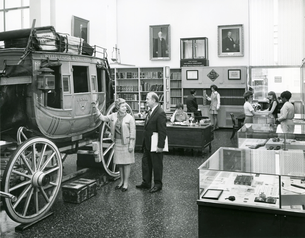 A large room with display cases along the back wall and one in the foreground to the right. To the left a stagecoach is displayed. A man and a woman stand talking before it. Image link enlarges image.